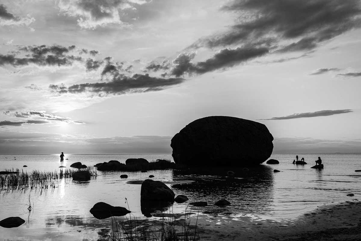 Černobílá fotografie pobřeží s velkým balvanem v mělké vodě, siluetami lidí a klidnou hladinou při západu slunce.