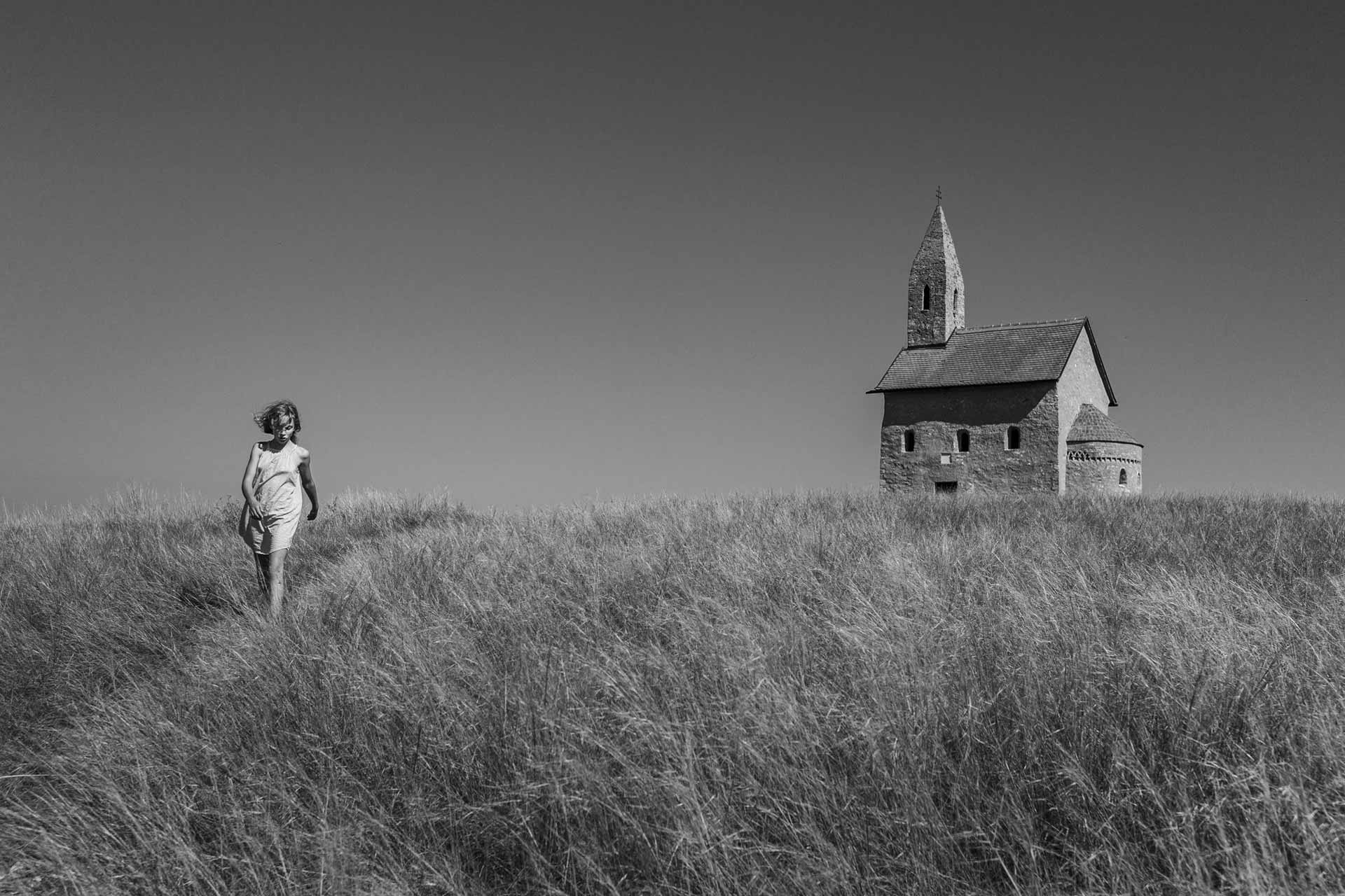 Dívka kráčí vysokou trávou, v pozadí stojí malý kostel.