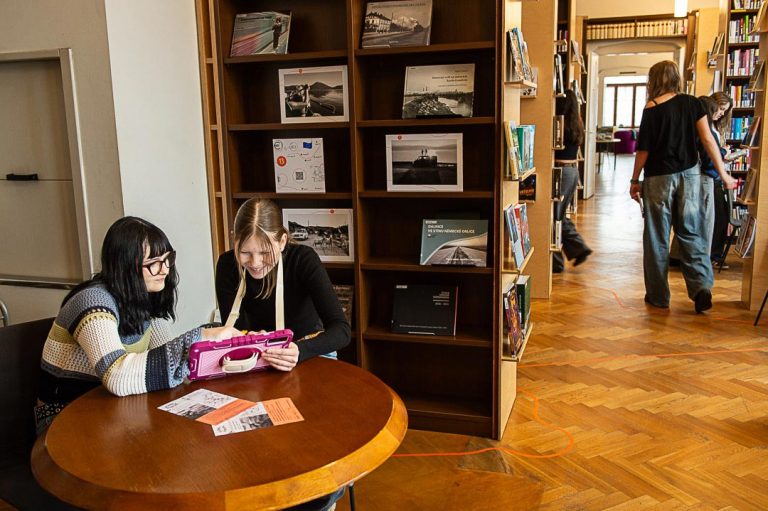 Dvě dívky sedí u kulatého stolu v knihovně a soustředěně sledují tablet položený na stole, kolem nich jsou knihy a informační letáky.