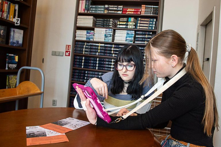 Detail dvou dívek sedících u stolu v knihovně, které si navzájem ukazují něco na tabletu a diskutují nad jeho obsahem.