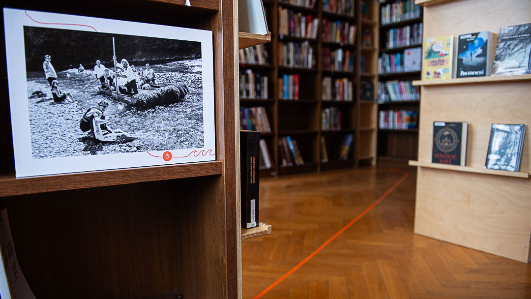 Černobílá historická fotografie vystavená na dřevěném panelu v knihovně, v pozadí jsou regály s knihami.